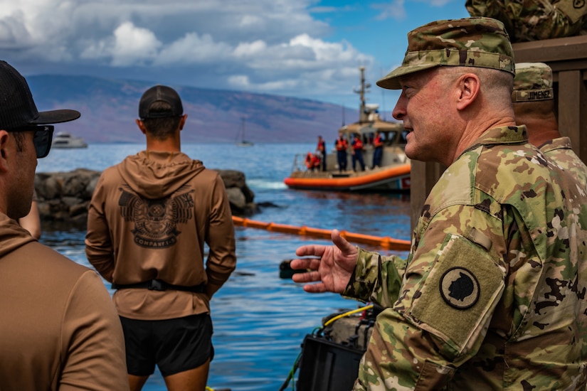 A man in uniform speaks to service members near a body of water.