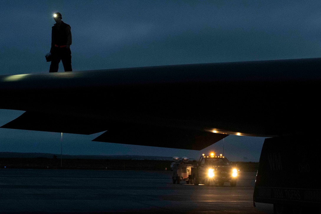 An airman shown in silhouette walks on the wing of a parked aircraft at night.