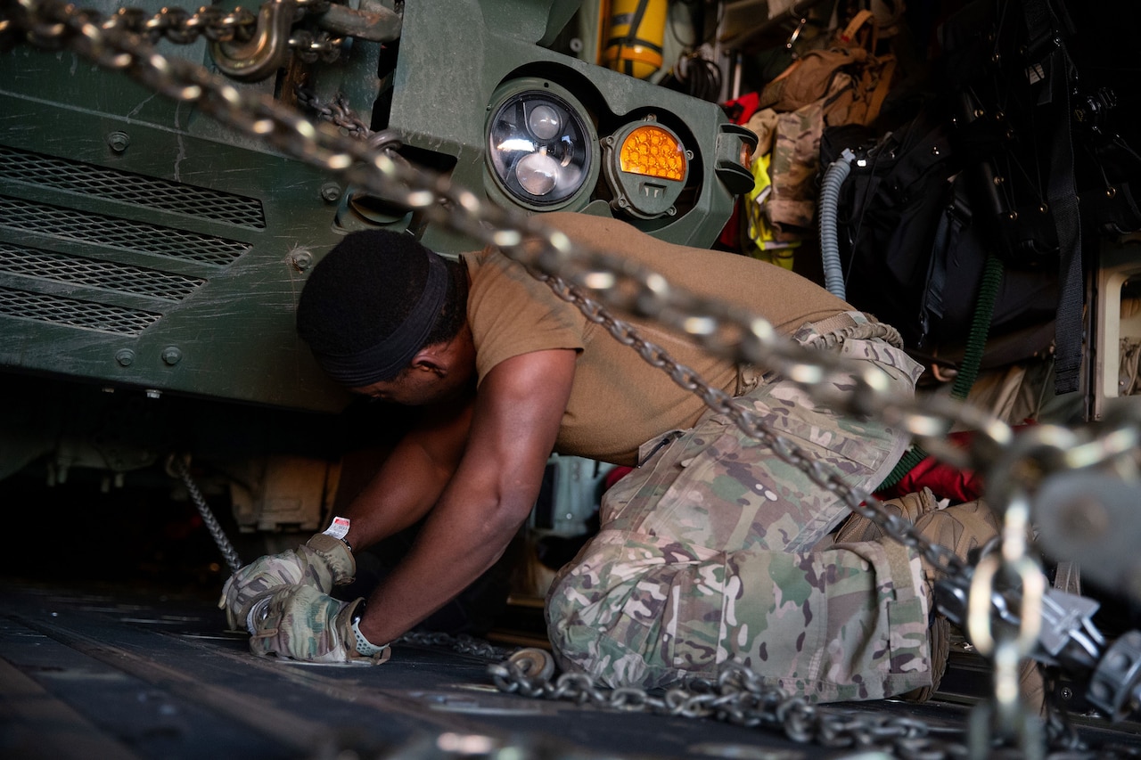 A person in military uniform pulls a chain under a military vehicle.