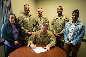 Five men in uniform, with one sitting and signing a paper is joined by two women for a group photo