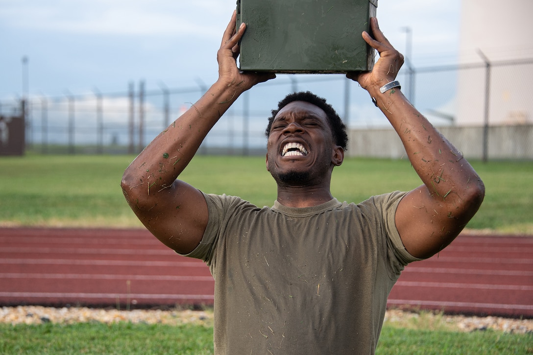 A service member winces while lifting a green metal box over his head.