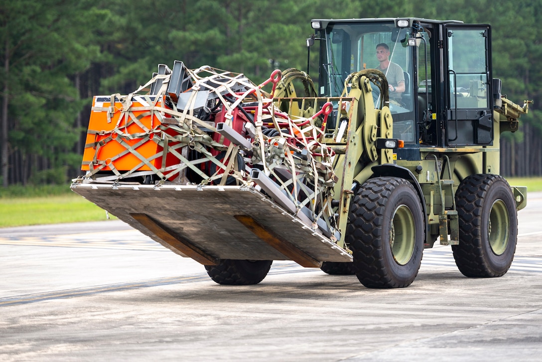 An airman operates a front-end wheel loader carrying supplies with a wooded background.