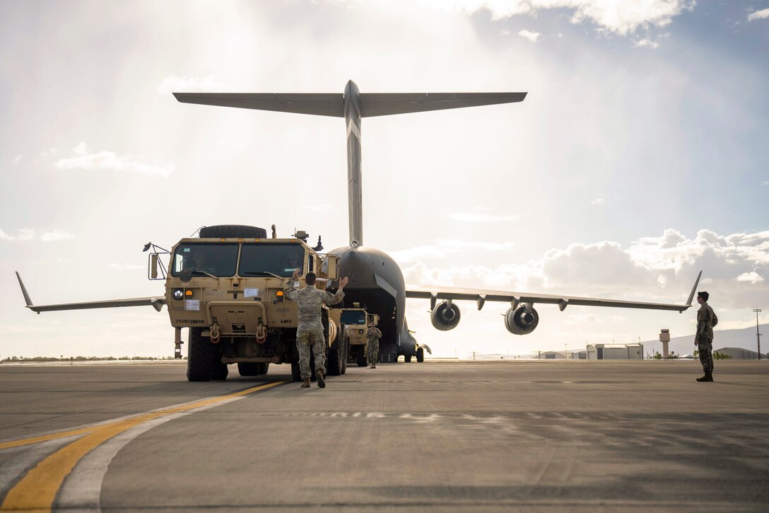 A service member watches from near the wing of a parked aircraft as another directs two vehicles into the back of the plane.