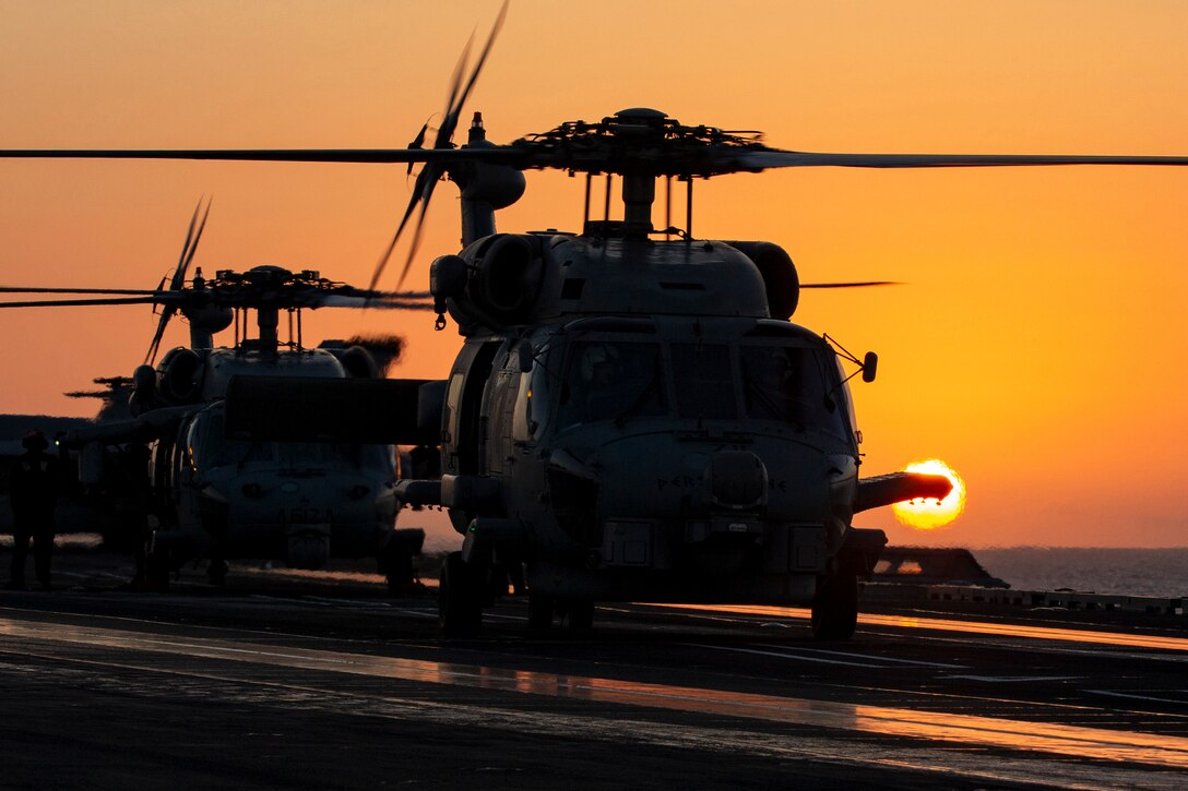 Two helicopters sit on the flight deck of a ship at twilight.