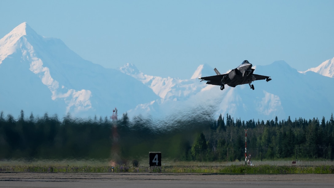In flight F-35A Lightning with Alaskan mountain range in the distance and runway in the foreground.