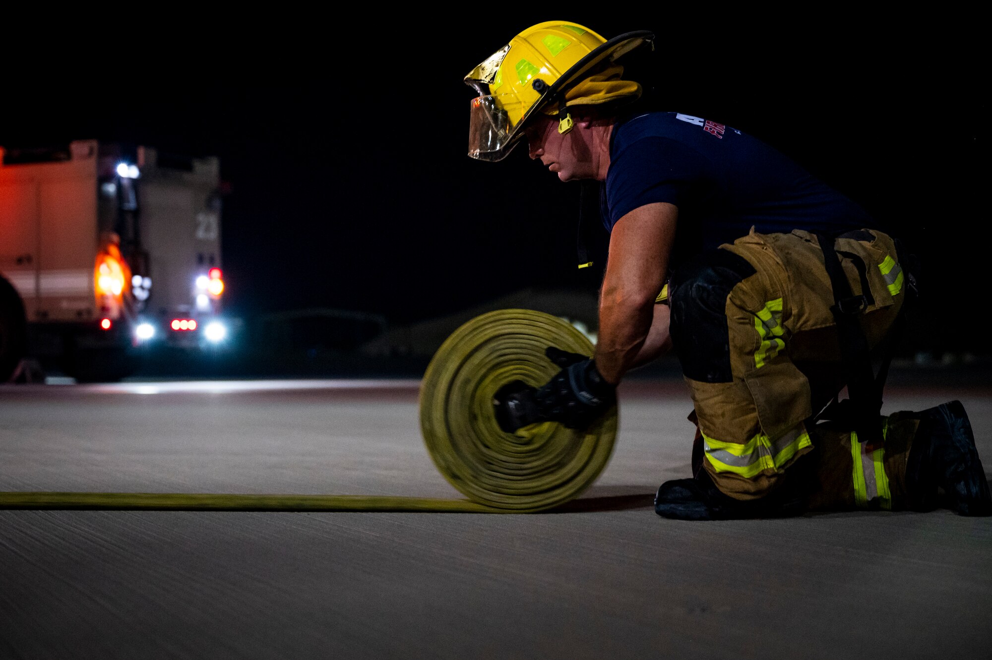 Military member wearing firefighting gear rolls up a fire hose.