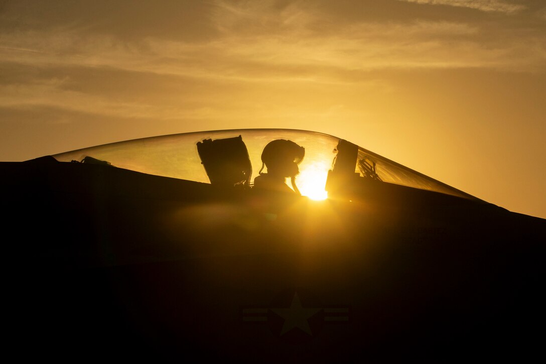 A pilot sits in the cockpit of a F/A-18E Super Hornet on the flight deck of a ship at twilight.