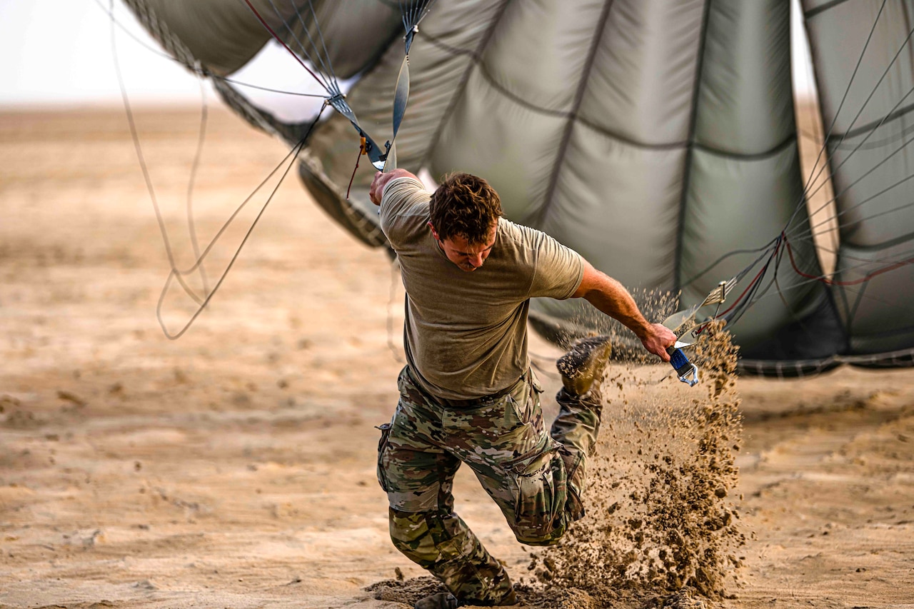 A soldier kicks up dirt while pulling on an open parachute.