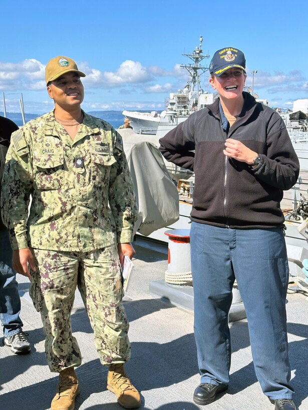 Photo is of a man and woman, both in Navy military uniform, standing on a the deck of a Navy ship