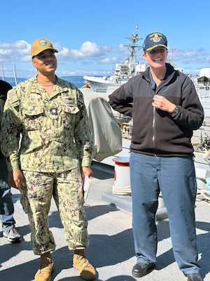 Photo is of a man and woman, both in Navy military uniform, standing on a the deck of a Navy ship