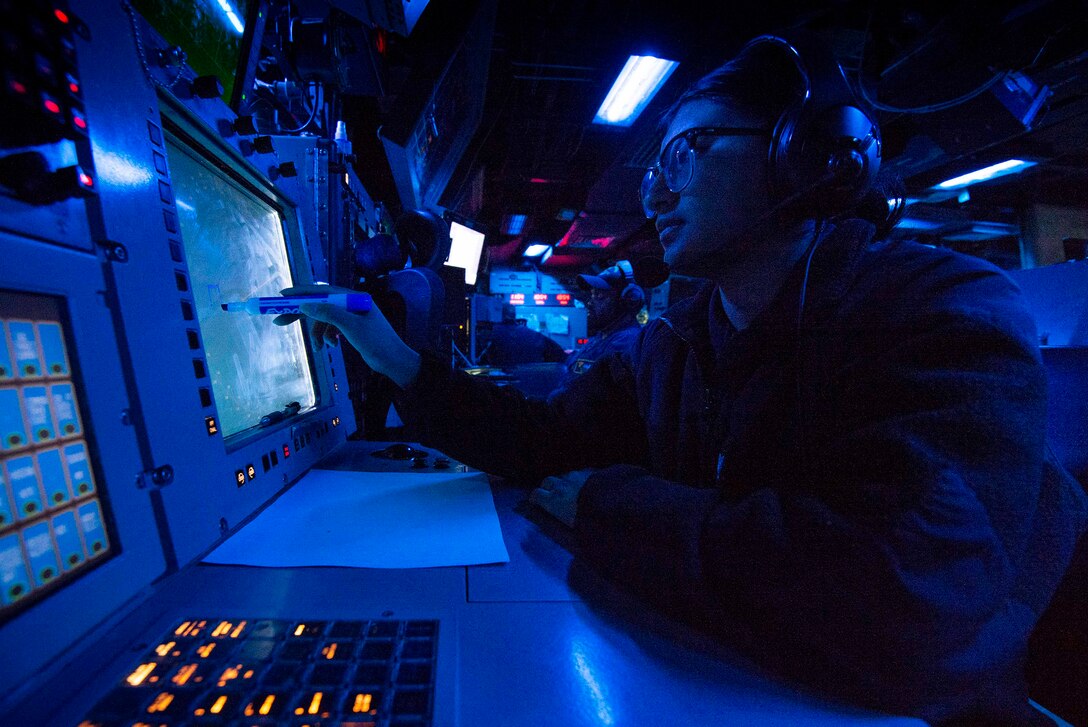 A sailor seated in a control room marks points on a computer monitor with a marker. The sailor is illuminated by blue light.