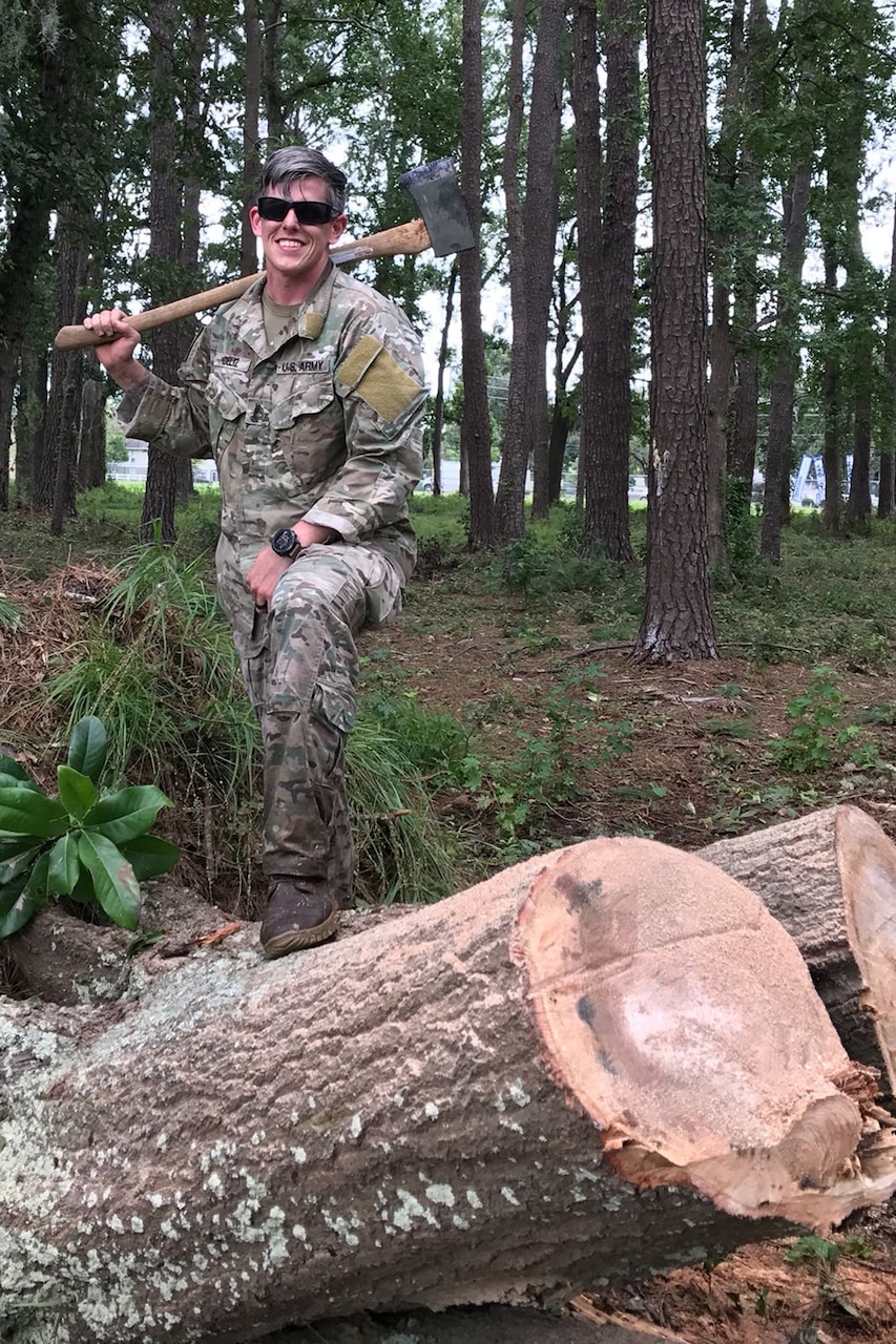A soldier holding an axe smiles as their leg leg is propped on a large log.