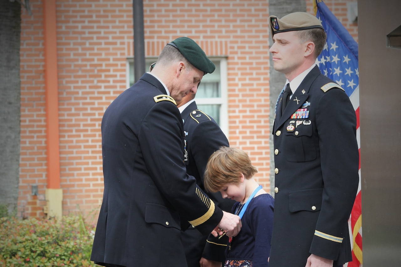 A child looks down on a medal a uniformed service member has placed around the child's neck. Two other uniformed service members stand beside the child.