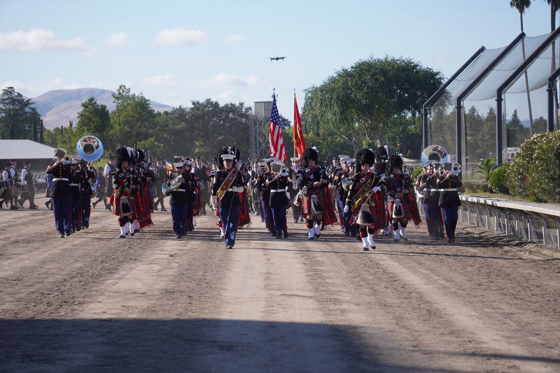 Two drum majors lead a combined Marine band and pipe band on the march down a dirt horseracing track. Behing them is a Marine color guard carrying the US Flag and Marine Corps flag.
