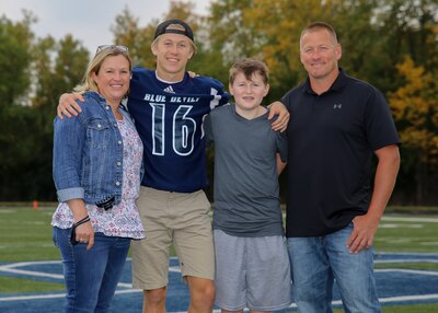 A family of four, mother, two sons, and a father stand with arms around each other. the son next to the mother is the tallest in the photo with the dad a close second. the younger son is the shortest. all are smiling at the camera. the photo looks to be taken outside at a football field with trees in the background.