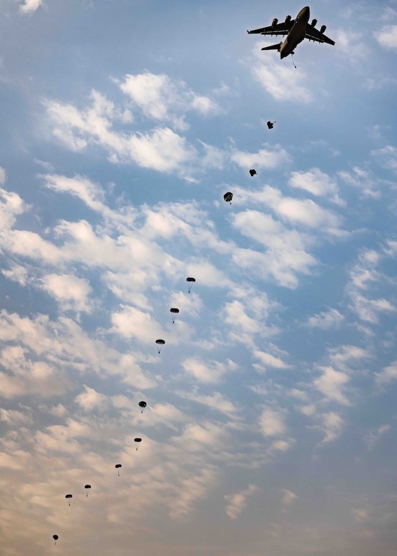 Service members descend in the sky wearing parachutes underneath an airborne aircraft,