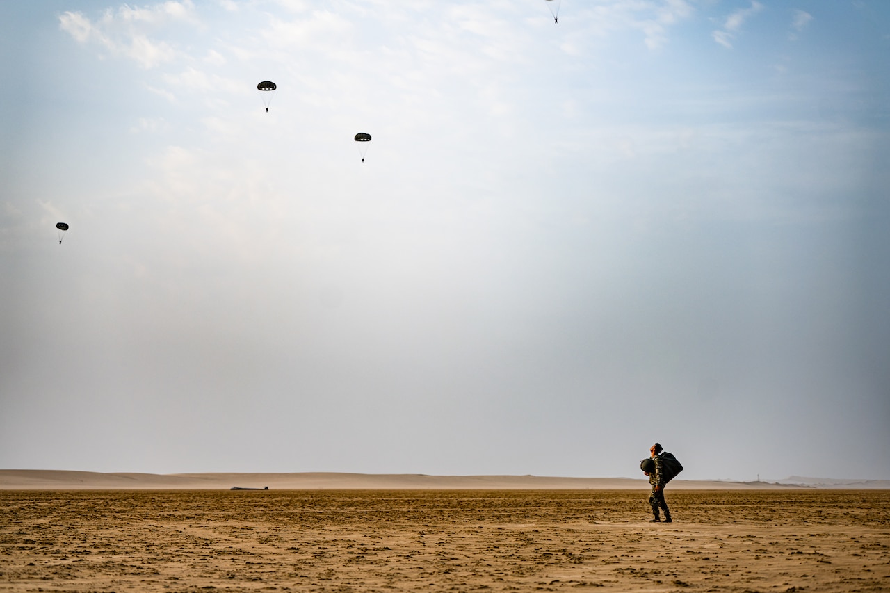 A soldier watches troops descend in the sky wearing parachutes.