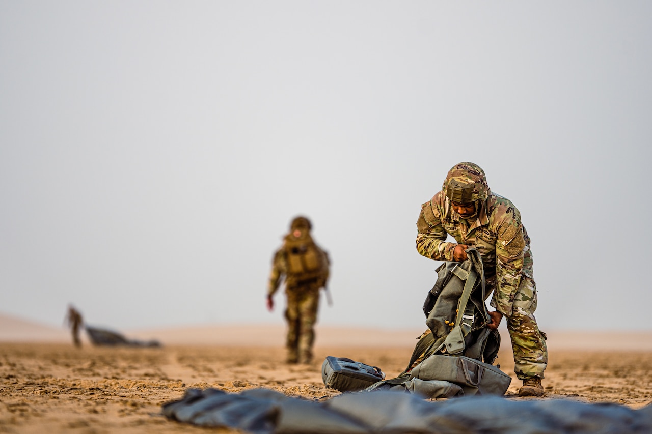 A soldier packs up a parachute as other soldiers walk behind.