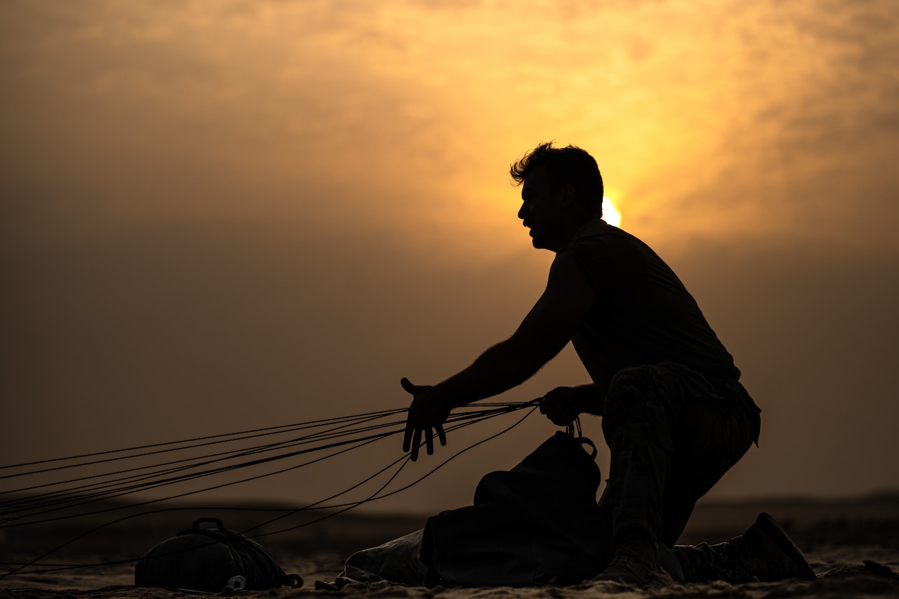 A soldier shown in silhouette pulls the ropes of a parachute.