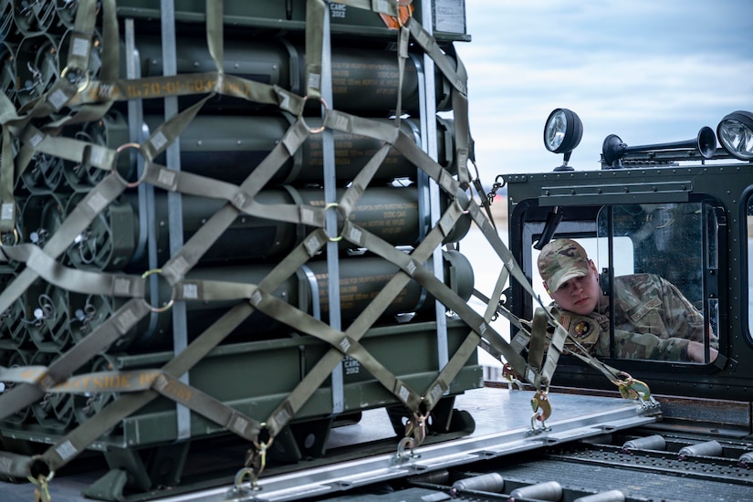 A service member operates a cargo loading vehicle carrying a pallet of munitions.