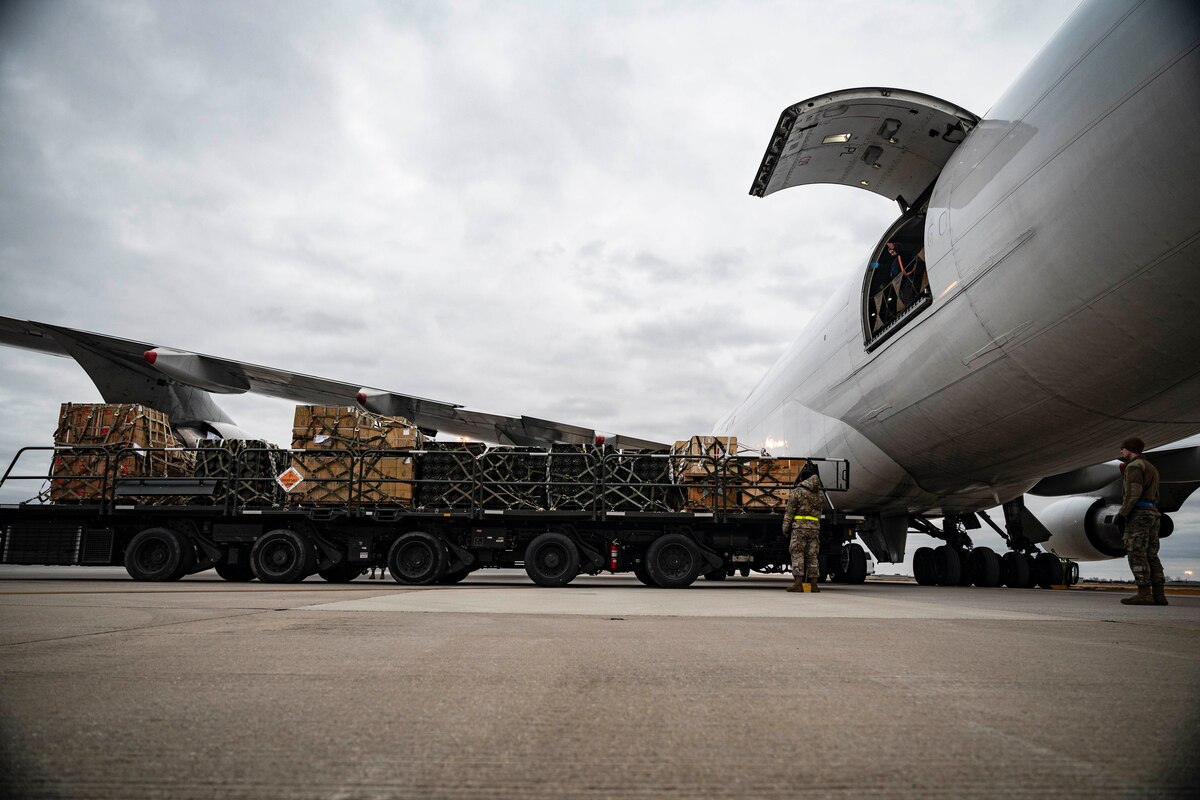 Service members stand near a cargo loading vehicle loaded with pallets of equipment near a transport airplane.