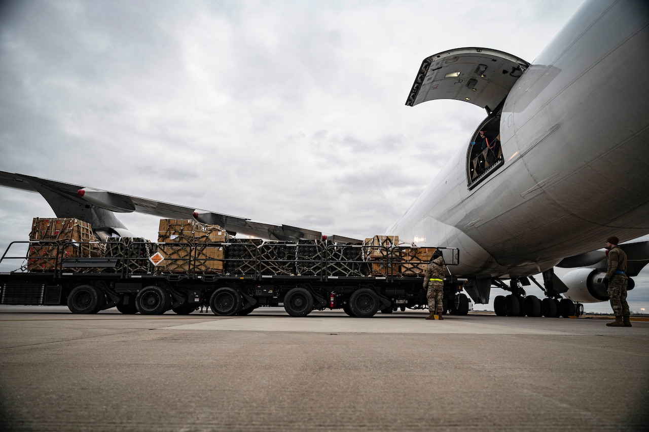 Service members stand near a cargo loading vehicle loaded with pallets of equipment near a transport airplane.
