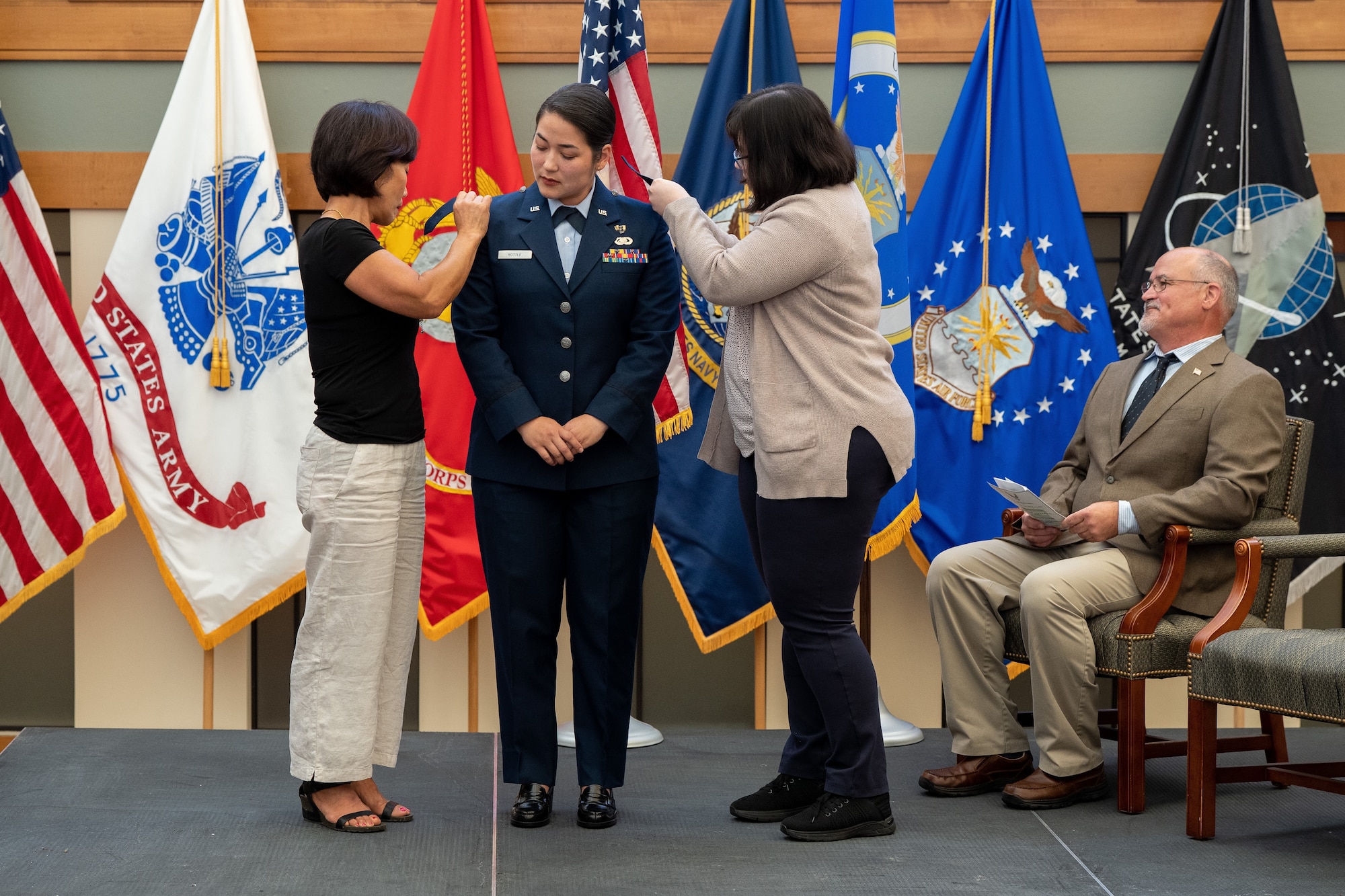 A woman stands on a stage while tow other women adjust her uniform.