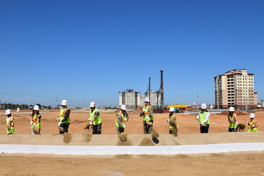 Ten people in safety vests and hard hats stand in a line throwing dirt with shovels. In the background is construction activity.