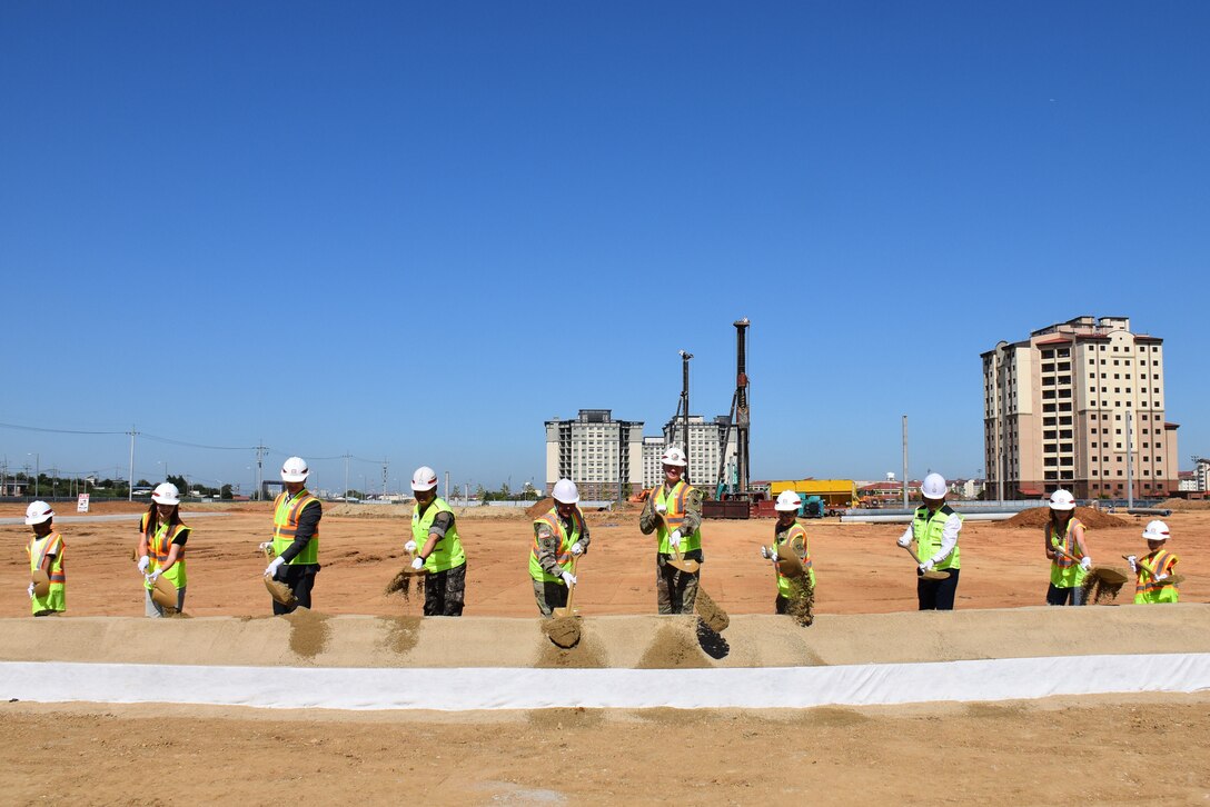 Ten people in safety vests and hard hats stand in a line throwing dirt with shovels. In the background is construction activity.