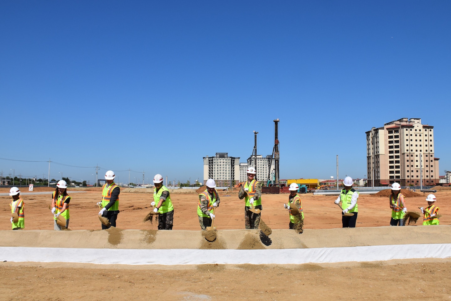 Ten people in safety vests and hard hats stand in a line throwing dirt with shovels. In the background is construction activity.
