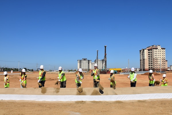 Ten people in safety vests and hard hats stand in a line throwing dirt with shovels. In the background is construction activity.