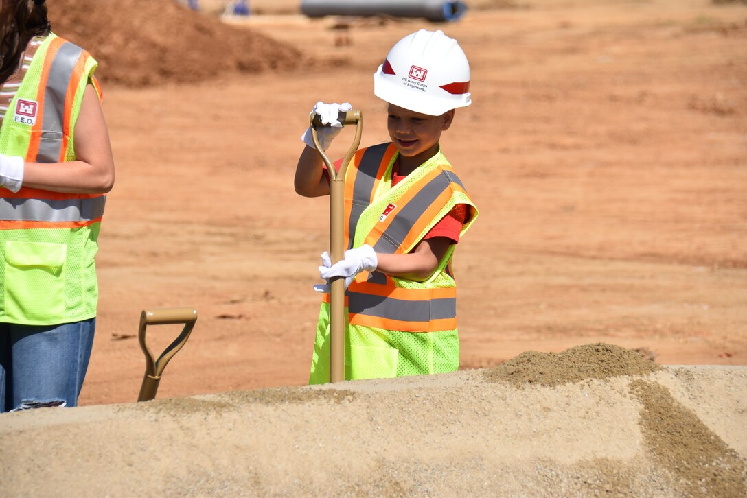 A young girl digs a shovel in dirt while wearing a safety vest and hard hat.