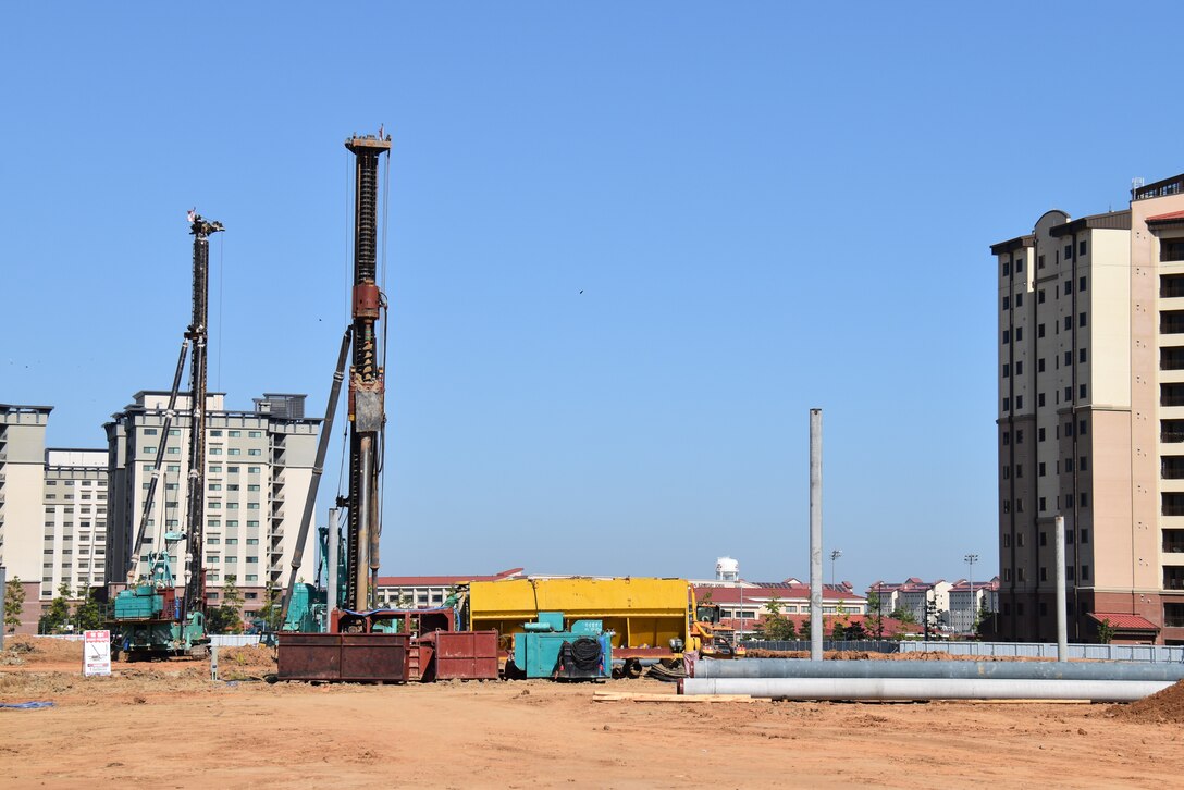 Pile drivers and other construction equipment sit on a construction site with housing towers in the background on Camp Humphreys.