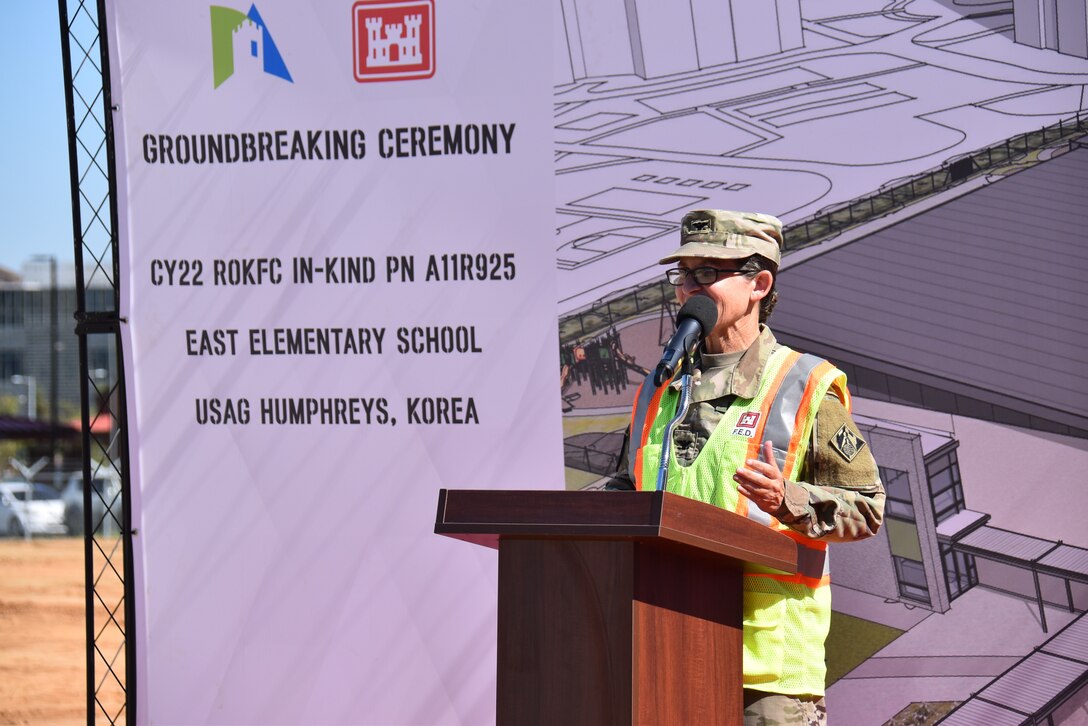 Col. Heather Levy, USACE FED commander, delivers remarks behind a brown podium while wearing a FED branded safety vest. She is in front of an artistic depiction of the finished elementary school.