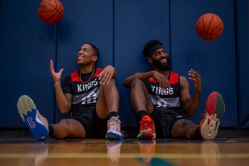Two men wearing basketball jerseys pose for a photo while tossing basketballs in the air and smiling.