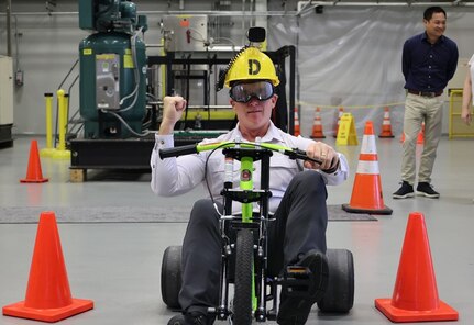 A man sits in a low tricycle riding between orange cones in a warehouse facility. The passenger is making a funny face and holing up his right arm in a fist. He dons a yellow hard hat with a camera and large goggles.