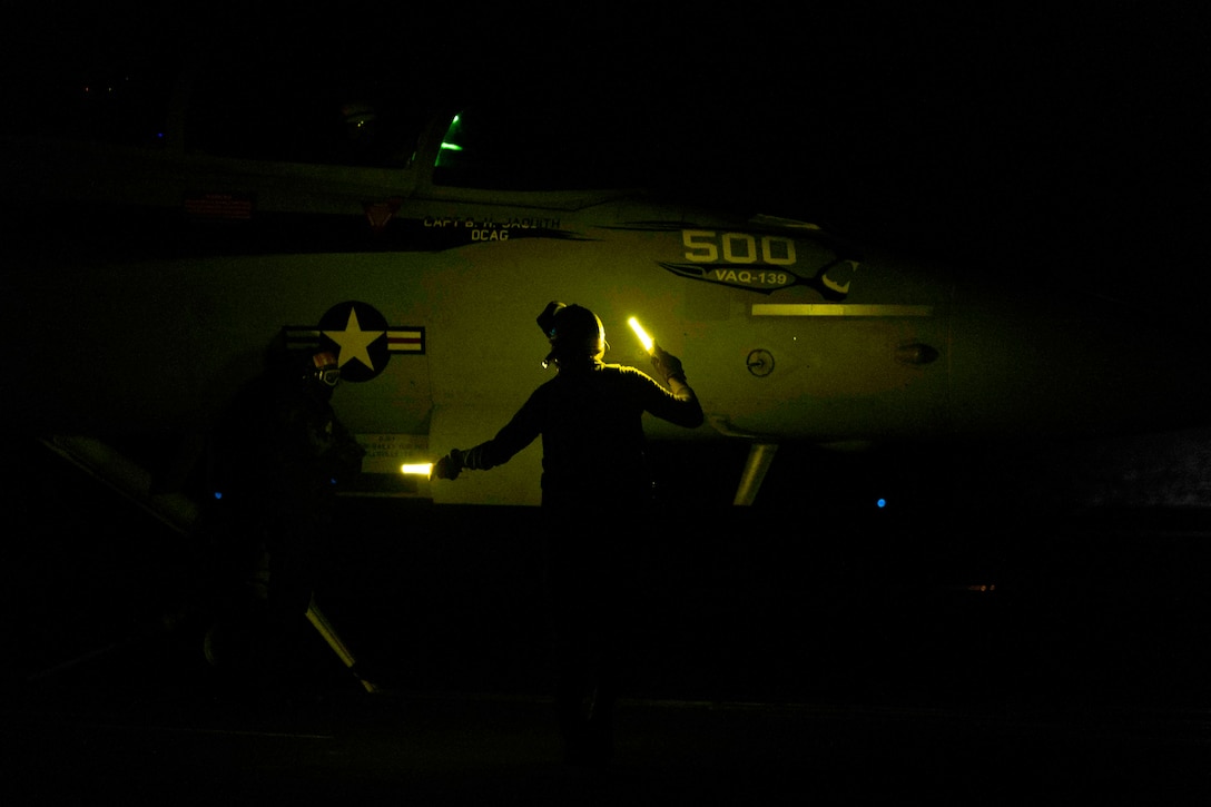 A sailor holding two flashlights inspects an aircraft at night.