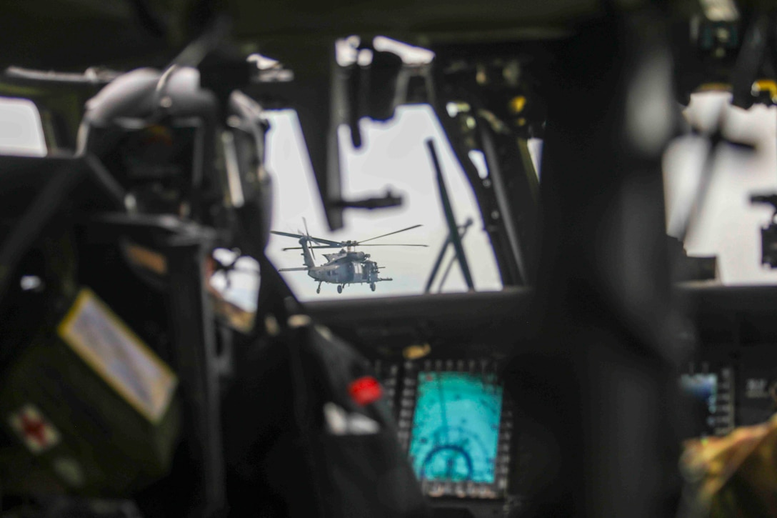 A soldier sits inside the cockpit of an airborne aircraft looking out toward another airborne aircraft.