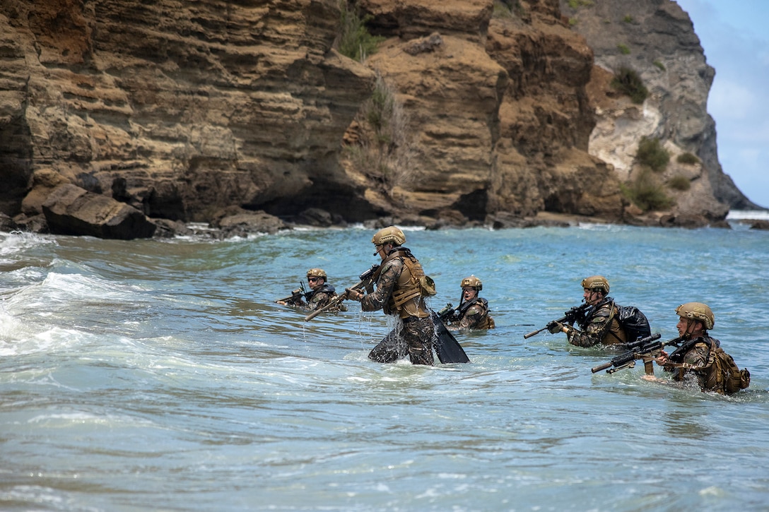 Uniformed Marines with weapons emerge from open water and walk toward the shore.