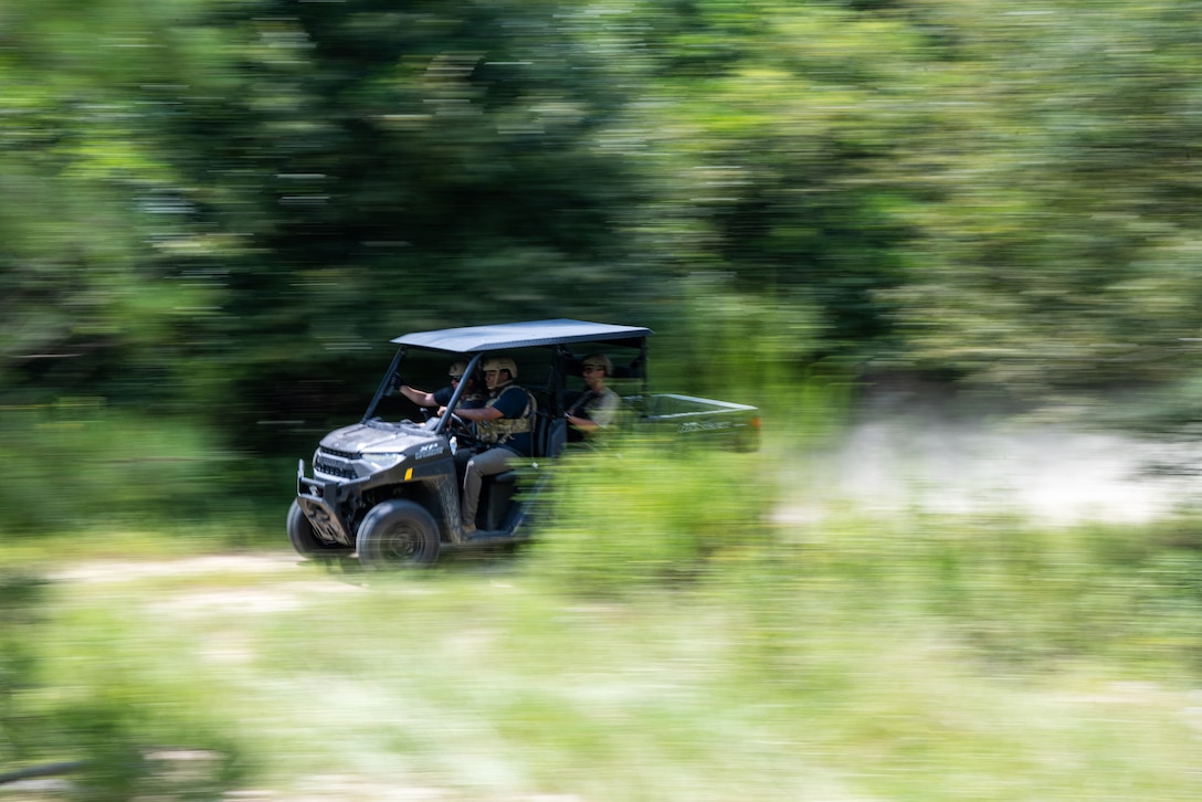 Airmen ride in a golf cart-like vehicle through a wooded area. The front of the cart is in focus with the surrounding areas blurred, indicating high speed.