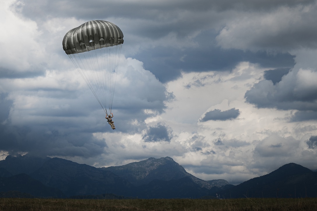 A soldier parachutes toward a field with mountains and a cloudy sky in the background.