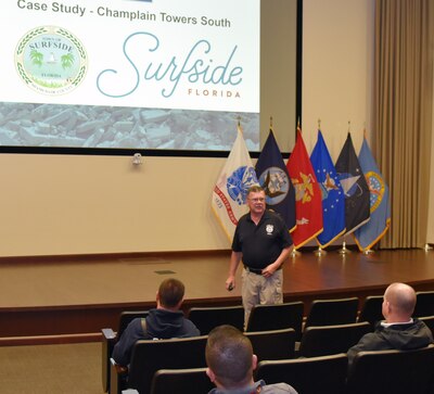 Photo is of a man standing in an auditorium talking to people.