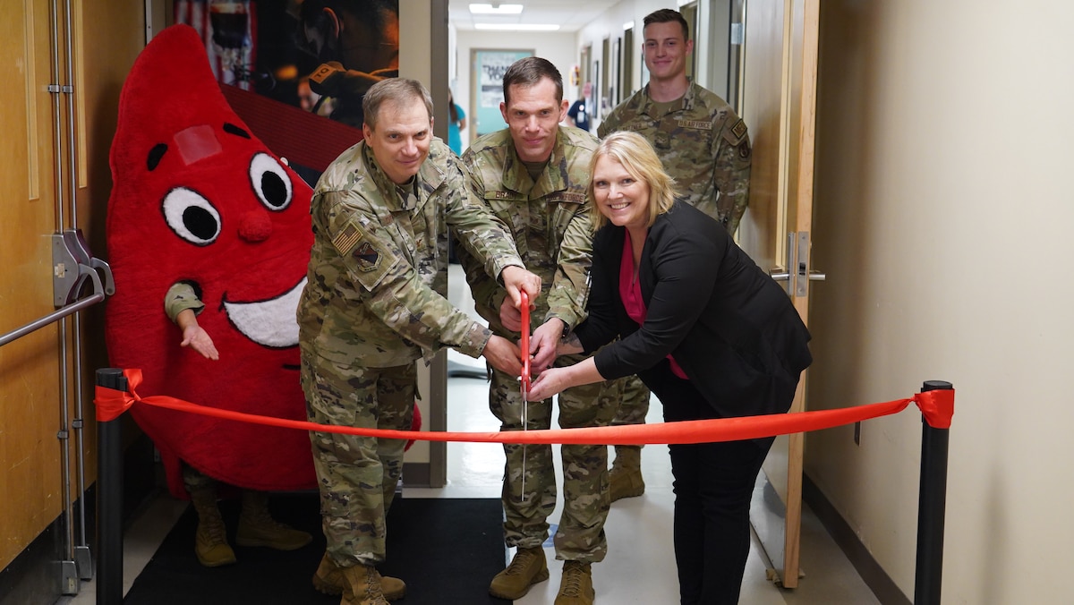 Two Airmen and a civilian cut a red ribbon with big scissors. A blood drop mascot in the background.