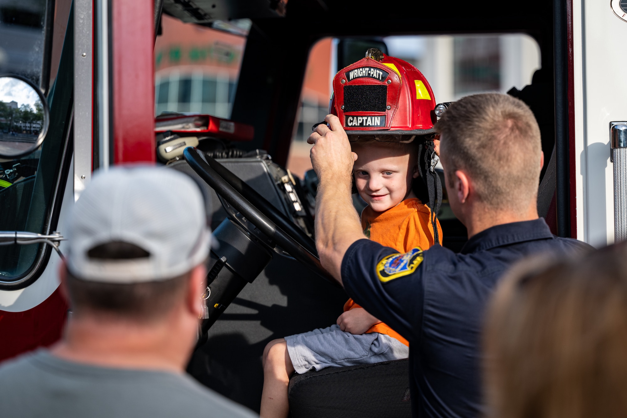 A young boy sits in the cap of a fire truck as a man in a firefighter's uniform, places a red fire helmet on his head.