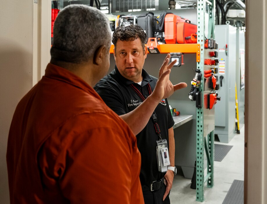 A man in a firefighter uniform, facing the camera, talks to a man with his back to the camera. The men are standing in a doorway with a machinery space in the background.