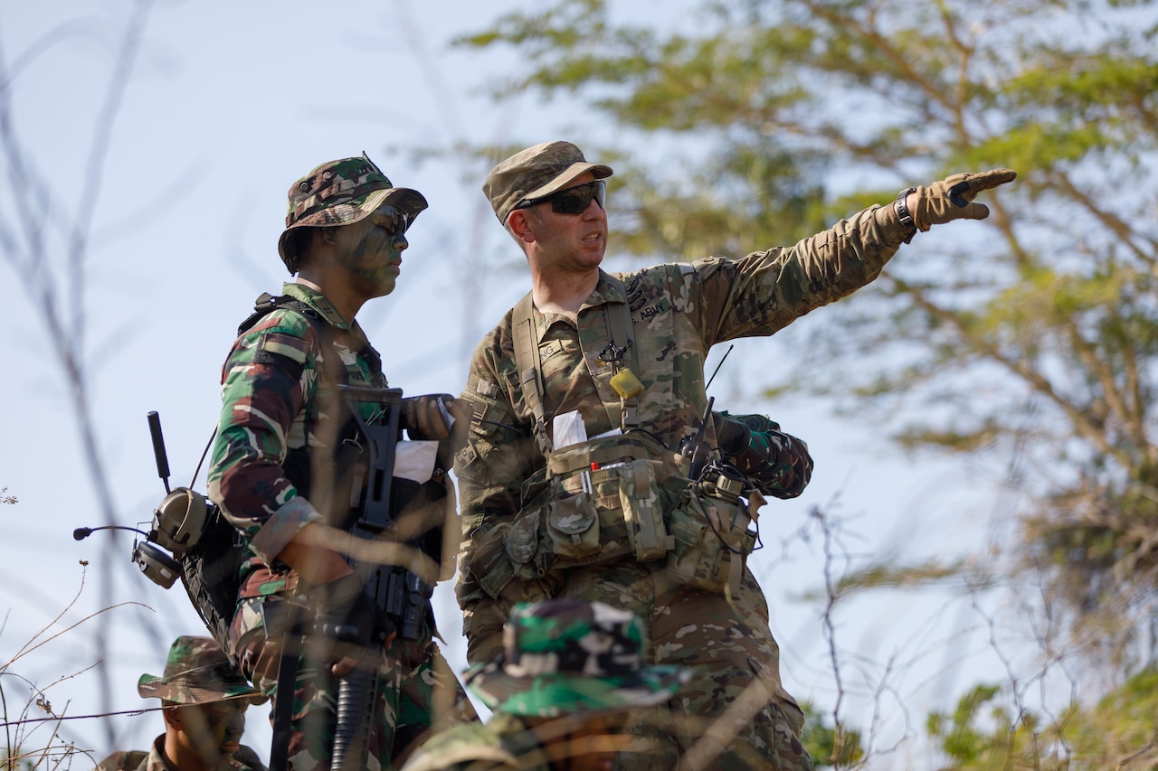 A U.S. soldier in battle gear points while standing next to another soldier in a field.