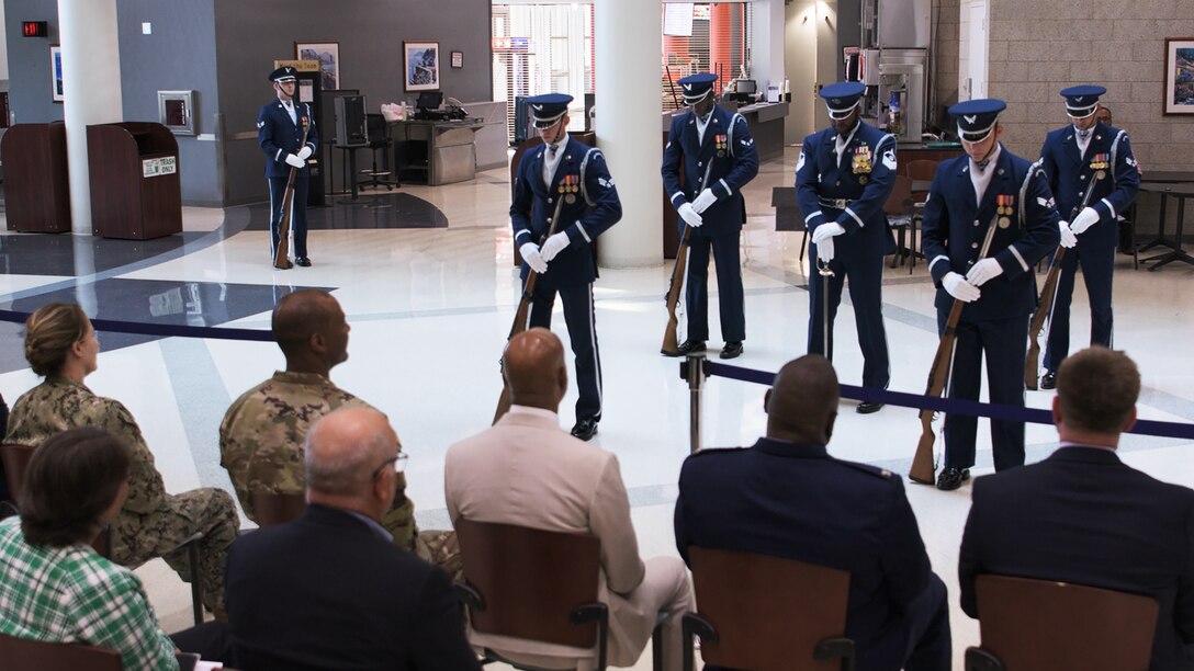 Spectators watch as men in dress Air Force uniforms and rifles stand at parade rest before beginning a performance.