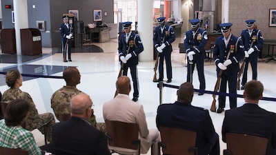 Spectators watch as men in dress Air Force uniforms and rifles stand at parade rest before beginning a performance.