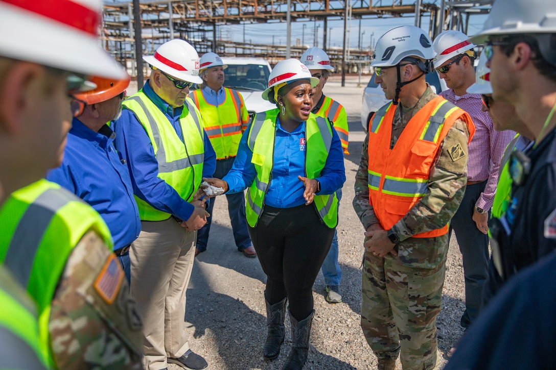 people in hard hats and safety vests are standing in a large group in an oil refinery, talking.