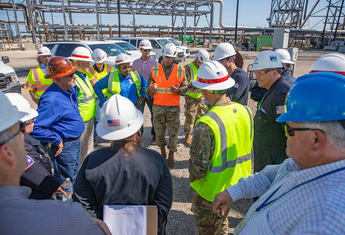 people in hard hats and safety vests are standing in a large group in an oil refinery, talking.